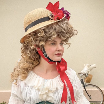 Woman wearing the Hair Helmet 1789, a bicycle helmet with a curly wig-style cover and straw hat adorned with red and purple flowers, styled with a ruffled dress and green corset